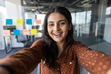 Smiling woman takes selfie in contemporary office setting. Bright sticky notes on glass wall highlight collaboration. Wood-colored attire contrasts sleek workspace. Optimistic and engaging atmosphereの写真素材