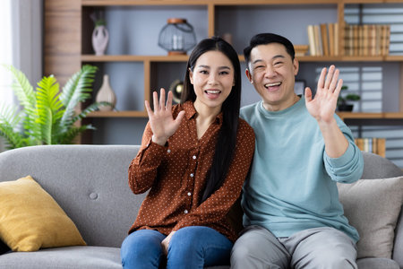 Happy asian couple sits on couch, smiling and waving at camera, conveying warmth and joy in modern living room. Perfect depiction of connection, family, and harmony.の写真素材