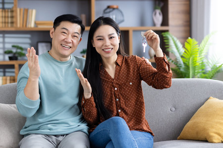 Asian couple joyfully presents new home keys while sitting on sofa in stylish living room, celebrating new beginnings and future together.の写真素材