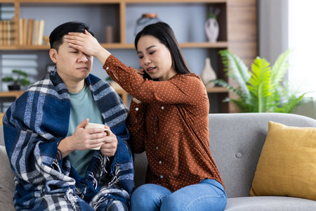Asian family scene showing woman comforting sick man on sofa. He holds cup, covered in blanket. Woman checks his temperature with care and concern, creating warm atmosphere.の写真素材