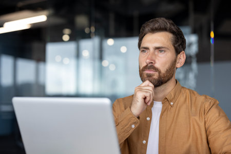 Man in casual shirt thinking while using laptop in modern office setting, reflecting on work ideas. Natural light filters through glass walls, creating focused atmosphere.の写真素材