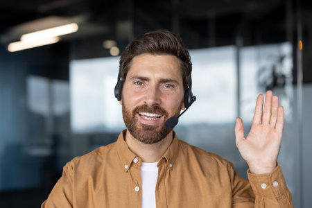 Confident man in office wears headset, smiling warmly while waving. Represents customer service, communication skills, professionalism. Ideal for business, support, tech, positive work environment.の写真素材