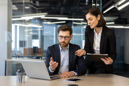 Colleagues in formal attire engaged in business meeting. Woman pointing at laptop screen, man contemplating solution. Office desk with laptop, smartphone, and documents enhancing collaborativeの写真素材