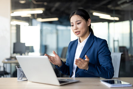 Asian business woman in modern office setting engaged in video conference using laptop. Expressive gestures suggest active communication and engagement. Professional environment indicates focusの写真素材