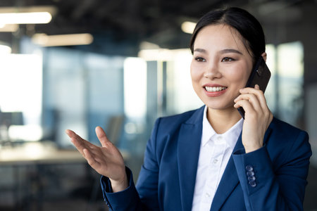 Asian businesswoman in blue suit talks on smartphone, discussing work in office. Confident expression suggests professionalism, career ambition, effective business communication.の写真素材