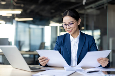 Asian businesswoman wearing glasses reviews documents at office desk. Laptop open, papers in hand. Professional setting emphasizes focus, organization, success. Modern office with natural light.の写真素材