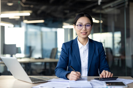 Confident Asian business woman in blue suit working at office desk with laptop and documents. Professional expression, focused on tasks, representing modern business environment.の写真素材