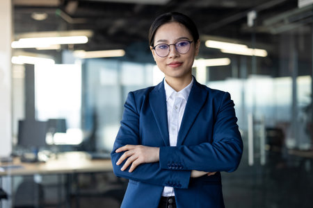 Asian business woman confidently standing in office, arms crossed, wearing glasses and suit. Emphasizes professionalism, leadership, corporate success, female empowerment in modern workplace.の写真素材