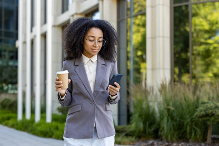 Confident African American businesswoman in gray suit checking smartphone while holding coffee cup outdoors. Image captures modern urban lifestyle, professional attire, and mobile technology.の写真素材