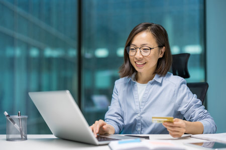 Asian businesswoman using laptop, holding credit card. Smiling, engaged in online shopping or electronic payment. Professional setting emphasizes modern business, finance, technology.の写真素材