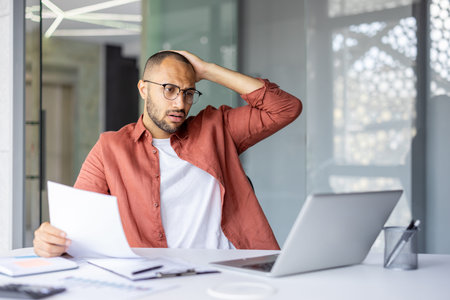 Worried man in office holding document while looking at laptop with stressed expression....