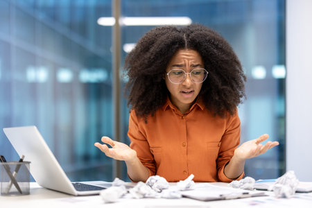 Frustrated woman in office surrounded by crumpled papers, laptop open, expressing confusion....