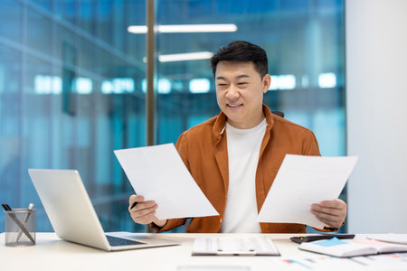 Smiling Asian businessman in casual attire reviewing documents at desk with laptop, symbolizing focus and satisfaction. Ideal for business themes, entrepreneurship, motivation.の写真素材