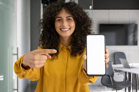 Smiling woman in yellow shirt points to blank smartphone screen in office. Concept of technology, presentation, business, communication, and digital display. Ideal for tech and marketing.の写真素材