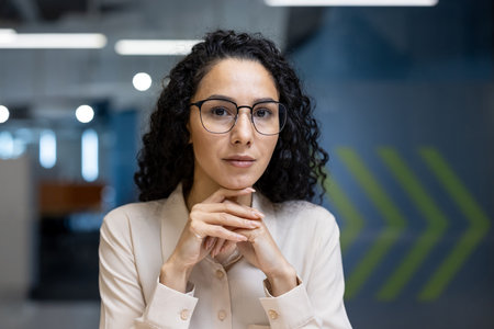 Confident businesswoman with curly hair wearing glasses in office setting, exuding professionalism and leadership. Her poised expression in professional attire communicates focus, determination.の写真素材