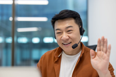 Asian man businessman in video call using headset for online meeting in modern office. Smiling, waving hand, showcasing remote communication, teamwork, technology for workplace connectivity.の写真素材