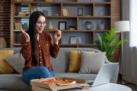 Asian woman with glasses sits on couch holding pizza slice while video chatting on laptop. She looks happy and surprised, enjoying leisure time in comfortable home environment.の写真素材
