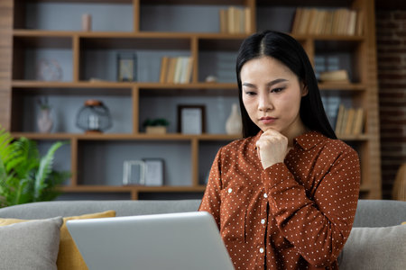 Thoughtful Asian woman using laptop at home, focusing intently on work or online learning. Comfortable setting enhances sense of concentration and productivity, surrounded by modern bookshelf.の写真素材