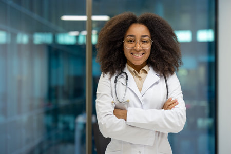 Confident African American woman doctor wearing white lab coat, standing with folded arms in...