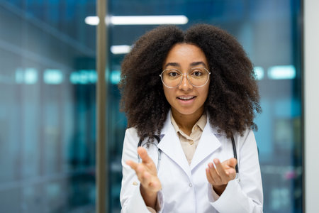 African American woman doctor wearing white coat in modern hospital, engaged in video call. Confident expression, looking at camera, conveying care, professionalism. Ideal for medical, technology concepts.の写真素材