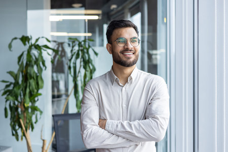 Confident young man in office standing by window, smiling and wearing glasses. Shows positivity,...