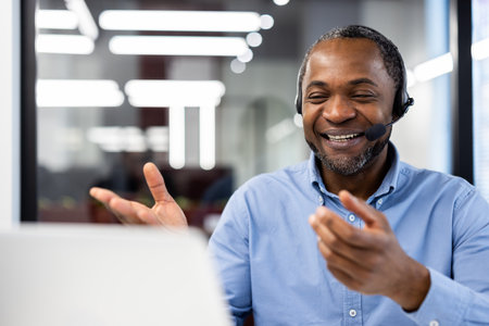 Smiling African American businessman wearing headset gestures during online meeting. Professional environment with focus on business communication. Mature man, confident, engaged, technology friendlyの写真素材