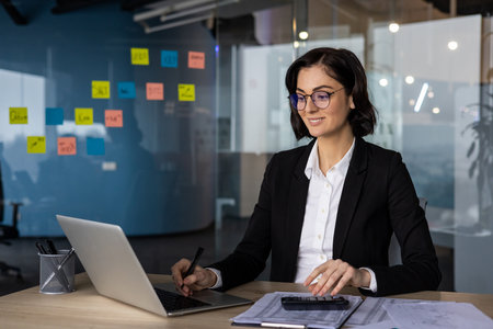 Businesswoman working in modern office, focused on laptop. Professional setting with sticky notes on wall, documents on desk. Office environment, creative planning concept.の写真素材