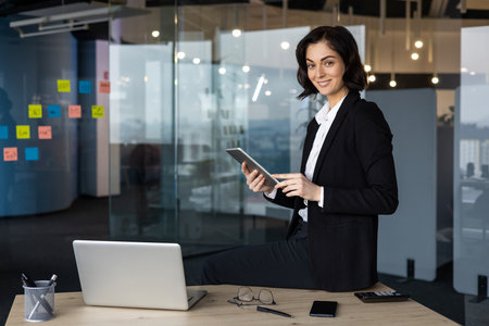Confident businesswoman in office using tablet, smiling at camera. Surrounded by work essentials like laptop, phone, and notes on glass wall. Concept of professionalism, technology, success.の写真素材