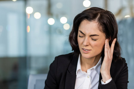 Woman at work experiencing ear pain, showing expression of discomfort. Dressed in formal business attire in office setting. Image conveys stress, possible hearing trouble, and workplace tension.の写真素材