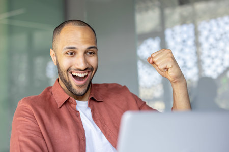Smiling young man with joyful expression celebrates success while working on his laptop in an office...