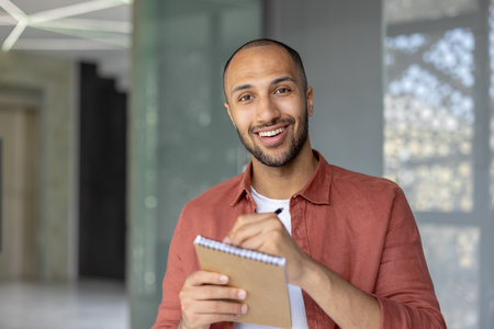A confident man with a friendly smile is holding a notepad and pen inside a modern space, showcasing a positive and productive atmosphere.の写真素材