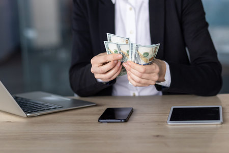 Confident businesswoman counts cash at office desk featuring laptop, emphasizing financial success,...