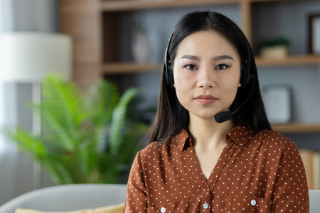 Asian woman wearing headset sitting in home setting, engaged in online communication. Ideal representation of remote work, tech interaction, customer support. Calm, professional demeanor.の写真素材