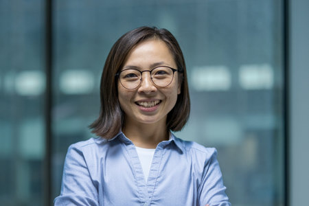 Professional portrait of a confident Asian woman in business attire wearing glasses, standing in a...