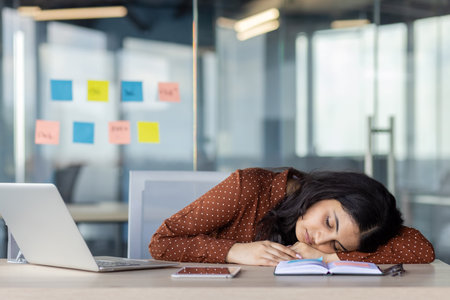 A young woman appears asleep at her office desk, resting on an open notebook beside a laptop and smartphone, illustrating exhaustion or a need for rest amidst a busy workplace setting.の写真素材
