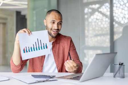 Smiling professional explaining statistical data while holding a chart and using a laptop, showcasing modern business communication.の写真素材