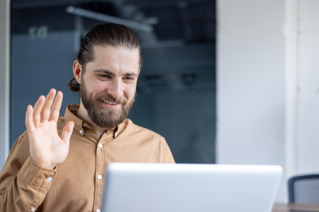 A bearded man in a casual shirt participates in a video call, waving and smiling warmly at the screen. Ideal representation for communication, remote work, or online professional interactions.の写真素材