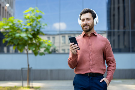 Successful businessman wearing headphones and with phone in hand walks through the business center of the city, office worker happily watches online video, dressed in red shirt.の写真素材