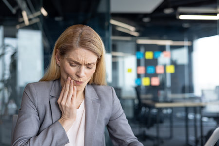 Mature woman businesswoman holding her jaw in pain at work in an office. Depicting discomfort possibly from a dental issue, this conveys feelings of distress and the need for healthcare attention.の写真素材