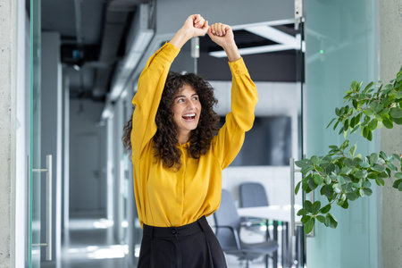 A joyful young woman celebrates with raised arms in a bright office. Her expression conveys excitement and accomplishment, epitomizing workplace success and positive energy.の写真素材