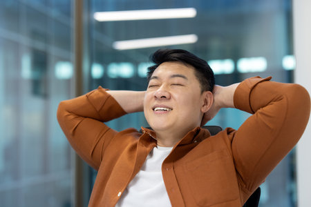 Asian man, dressed casually, leaning back with hands behind his head in modern workspace, expressing relaxation and satisfaction with a joyful smile.の写真素材