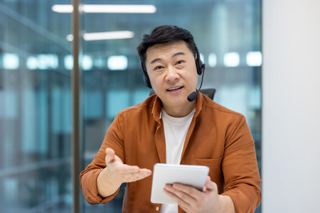 Asian man wearing a headset and holding a tablet while working in a call center, providing support to customers remotelyの写真素材