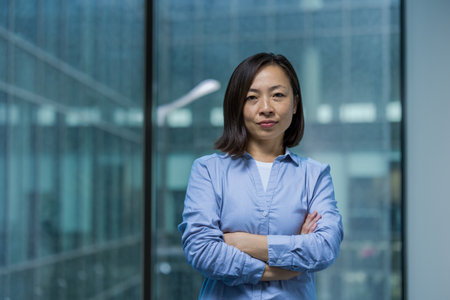 Portrait of an Asian businesswoman standing confidently with her arms crossed against the backdrop of a modern office building window.の写真素材
