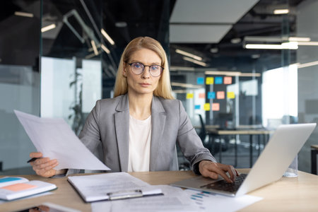 Businesswoman working at her desk with a laptop, documents, and wearing glasses in a modern office environment.の写真素材