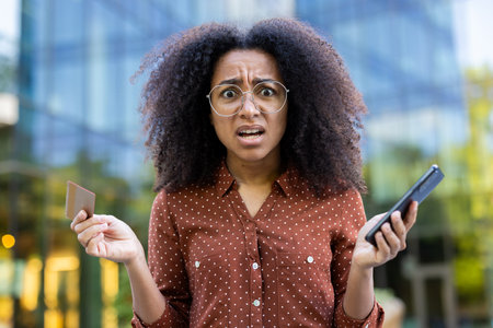 A shocked woman with glasses stands outdoors holding a credit card and smartphone, experiencing financial stress or fraud concern.の写真素材