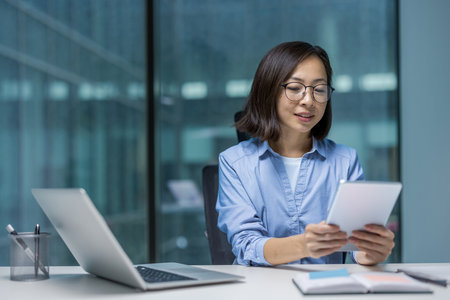 Young adult Asian woman in glasses works on her tablet at her desk in an office. Laptop and notepad are also on the desk.の写真素材