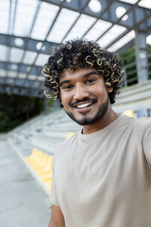 A young man smiles into the camera while video calling at the stadium, in a casual setting with...