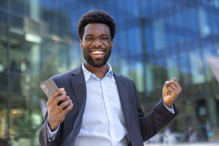 A cheerful businessman in a suit pumps his fist in excitement while holding his phone in front of a modern glass building, smiling widely.の写真素材