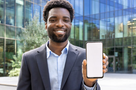 A cheerful African American businessman in a suit presents a smartphone with a blank white screen against a modern glass building.の写真素材