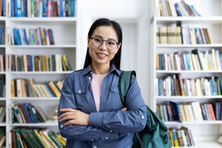 A smiling asian woman stands in a library, posing with her arms crossed, wearing glasses and holding a green backpack.の写真素材